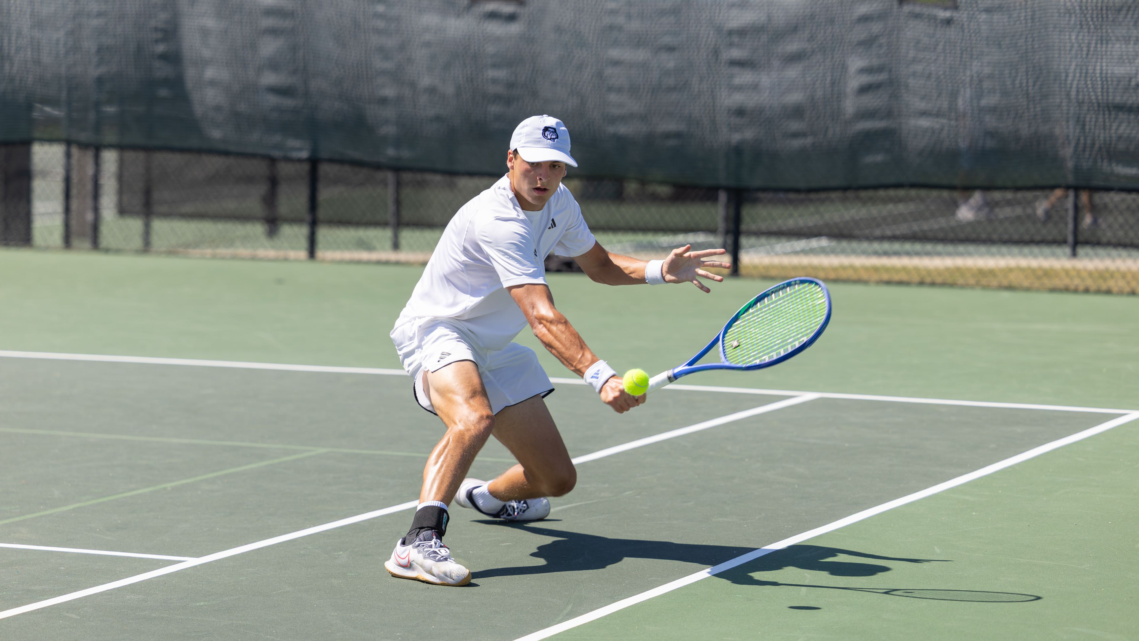 Trevor in a white tennis kit reaching forward to play a forehand.